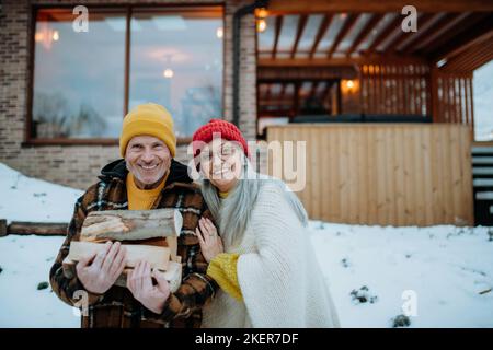 Portait des älteren Ehepaares, das Holzstämme trägt und das Feuer im Freien vorbereitet, während des Wintertages. Stockfoto