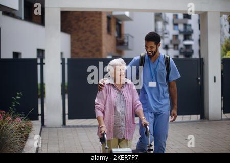 Pfleger, der mit einer älteren Klientin vor dem Pflegehaus läuft. Stockfoto