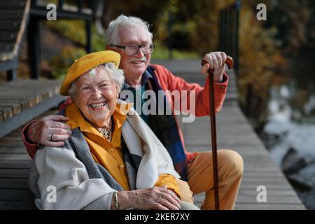 Glückliches Seniorenpaar in Herbstkleidung, das nach dem Spaziergang in der Nähe des Sees Pause gemacht hat. Stockfoto