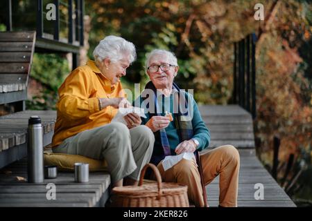 Glückliches Seniorenpaar in Herbstkleidung, das nach dem Spaziergang in der Nähe des Sees Picknick macht. Stockfoto