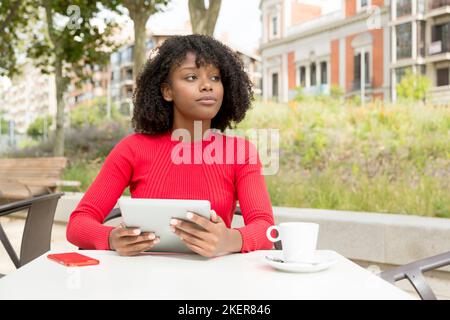 Nachdenkliche junge Frau, die mit einem Tablet arbeitet und Kaffee im Freien trinkt Stockfoto