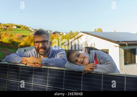 Vater mit seiner kleinen Tochter in der Nähe ihres Hauses mit Sonnenkollektoren. Alternative Energie, Ressourcenschonung und nachhaltiges Lifestyle-Konzept. Stockfoto