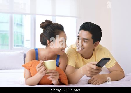 Glückliches junges vietnamesisches Paar mit einer Tasse Kaffee und einer Fernbedienung im Bett Stockfoto