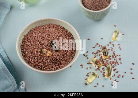 Leinsamen und gemahlene oder zerkleinerte Leinsamen in einer kleinen Schüssel braun und Gelatinekapseln mit Omega-Öl auf hellblauem Hintergrund. Stockfoto