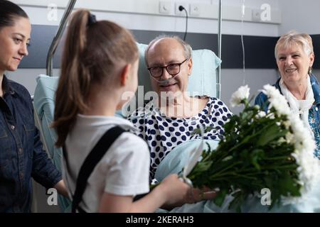Kranker Großvater erhält Blumen von Enkelin im Krankenhauszimmer. Bettlägeriger alter Mann, der von Verwandten im geriatrischen Klinikraum besucht wird. Kleines Mädchen, das einem bettlägerigen älteren Mann Blumen schenkt. Stockfoto