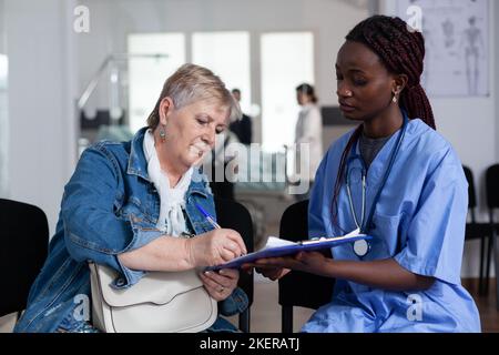 Ältere kaukasische Frau, die Entlassungspapiere im Bereich des geriatrischen Krankenhausausgangs unterschrieb. Afro-Krankenschwester, die eine alte Dame an der Klinikrezeption besucht. Leitender Patient, der medizinische Dokumente ausfüllt. Stockfoto