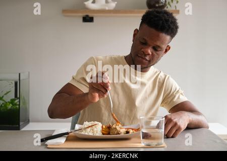 Junger afrikanischer Mann, der gebratenes Huhn, Reis und Kochbanane mit Tomatensauce isst. Stockfoto