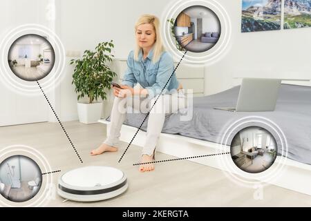 Glückliche junge Frau mit Smart-Home-Anwendung auf Smartphone und Roboter Staubsaugen Boden im Hintergrund im modernen Wohnzimmer. Stockfoto