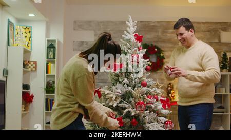 Paar mit passenden Kleidung schmücken ihre weihnachtsbaum für Winterferien. Dekorieren schönen Weihnachtsbaum mit Glaskugel Dekorationen. Frau und Mann in passenden Kleidern helfen verziertes Haus mit Girlande Lichter Stockfoto