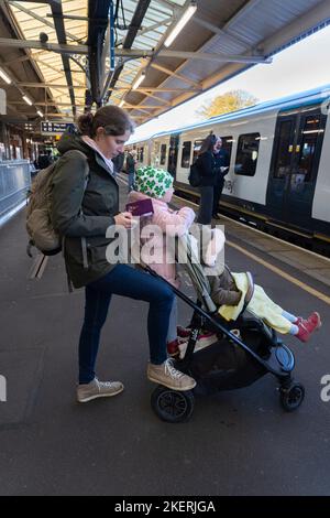 Eine Mutter, die mit ihren zwei kleinen Kindern und einem Kinderwagen auf ihrem Smartphone schreibt und am Bahnhof Basingstoke auf einen Zug wartet. England Stockfoto