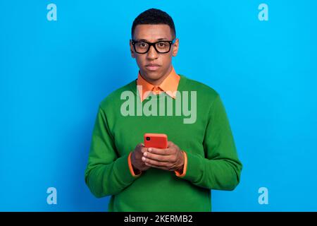 Foto von intelligenten klugen Mann tragen orange Hemd unter grünen Sweatshirt Eingabe Nachricht in Smartphone isoliert auf blauem Hintergrund Stockfoto