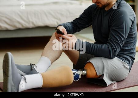 Nahaufnahme eines Mannes mit Beinprothese, der zu Hause auf einer Yogamatte sitzt und ein Smartphone verwendet Stockfoto