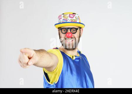 Urlaubskonzept. Ein Clown in einem leuchtend blauen und gelben Anzug, einer Brille und einem Hut zeigt seinen Finger nach vorne. Stockfoto
