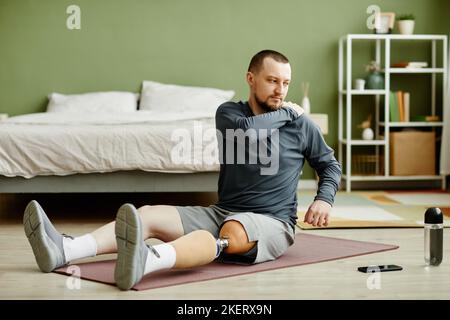 Ganzkörperportrait eines Mannes mit Beinprothese, der zu Hause Stretching-Übungen auf einer Yogamatte macht Stockfoto