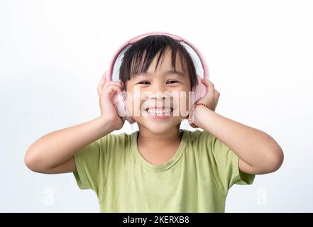 Glücklich lächelndes asiatisches kleines Mädchen, das Musik auf kabellosen Kopfhörern hört, die auf weißem Hintergrund isoliert sind. Stockfoto