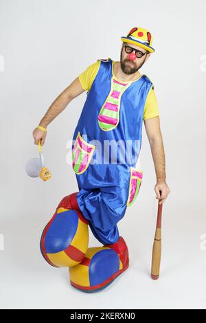Urlaubskonzept. Der Clown ist ein Mann in einem leuchtend blauen und gelben Anzug mit einem Baseballschläger und einem Lautsprecher. Stockfoto