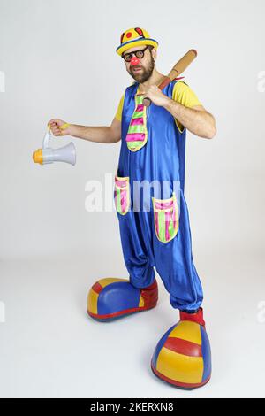 Urlaubskonzept. Der Clown ist ein Mann in einem leuchtend blauen und gelben Anzug mit einem Baseballschläger und einem Lautsprecher. Stockfoto