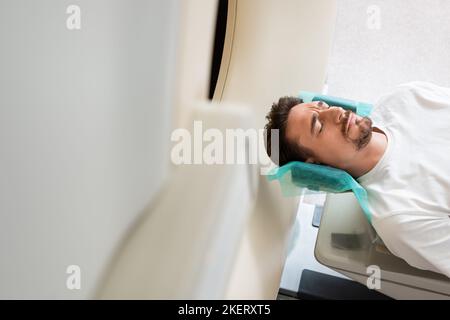 Besorgter Mann mit geschlossenen Augen während der Untersuchung auf CT-Scanner in der Klinik liegend, Stockbild Stockfoto