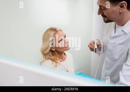Lächelnde blonde Frau, die einen positiven Arzt mit Ultraschallsonde in der Klinik ansieht, Stockbild Stockfoto