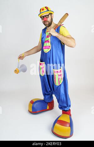 Urlaubskonzept. Der Clown ist ein Mann in einem leuchtend blauen und gelben Anzug mit einem Baseballschläger und einem Lautsprecher. Stockfoto