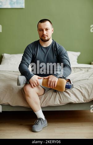 Vertikales Porträt eines Mannes, der ein prothetisches Bein hält, während er auf dem Bett sitzt und die Kamera anschaut Stockfoto