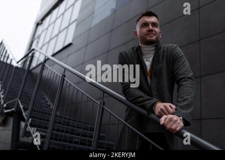 Ein starker brutaler Mann im Herbstmantel steht auf der Treppe und blickt in die Ferne Stockfoto