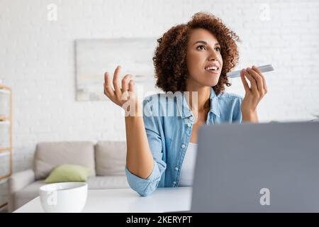 afroamerikanische Frau, die Sprachnachricht auf dem Smartphone in der Nähe von Laptop und Tee aufzeichnet, Stockbild Stockfoto