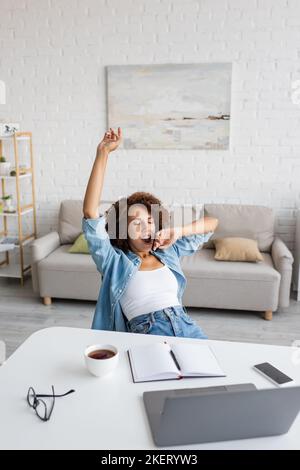 Müde afroamerikanische Frau mit geschlossenen Augen Gähnen und Stretching in der Nähe des Arbeitsplatzes zu Hause, Stock Bild Stockfoto