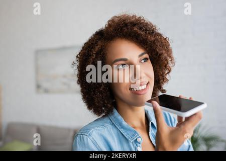 Fröhliche afroamerikanische Frau mit lockigen Haaren, die Sprachnachricht auf dem Smartphone aufzeichnet, Stockbild Stockfoto