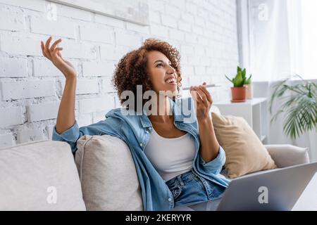 Glückliche afroamerikanische Frau, die Sprachnachricht auf dem Smartphone in der Nähe des Laptops aufzeichnet, während sie auf der Couch sitzt, Stockbild Stockfoto