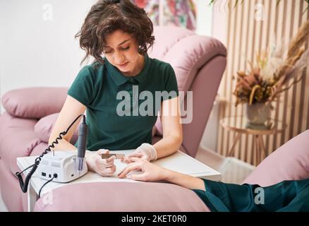 Manikurist Entfernen von Nagelhaut auf Client Hand mit elektrischen Nagelbohrer. Weibliche Maniküre Master sitzt am Tisch, während Hardware-Maniküre auf Frau Hand. Stockfoto