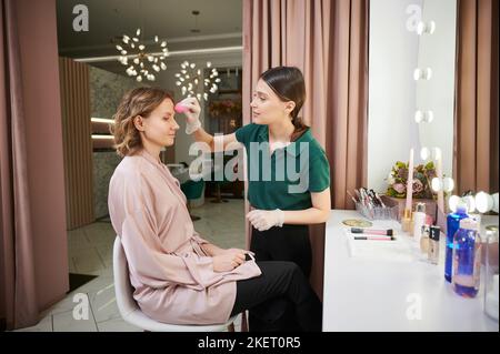 Junge Frau, die am Frisiertisch sitzt, während Make-up-Künstler professionelles Make-up macht. Schönheitsfachfrau, die das Fundament auf das Gesicht der Frau mit einem Blending-Schwamm aufgelegt hat. Stockfoto