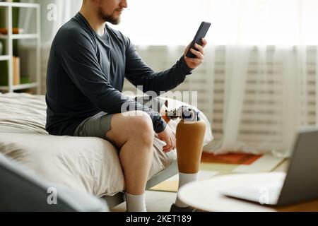 Seitenansicht Porträt eines Mannes mit Beinprothese mit Smartphone, während er zu Hause auf dem Bett sitzt Stockfoto