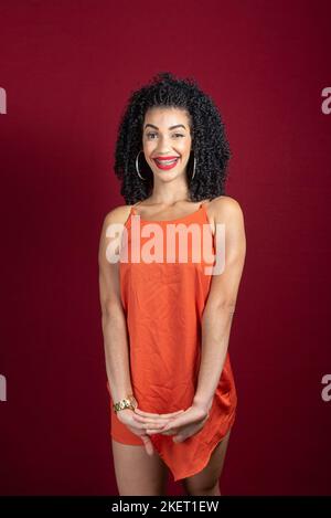 Junge, schöne lächelnde Frau, die im Studio mit orangefarbenem Outfit vor rotem Hintergrund steht. Stockfoto