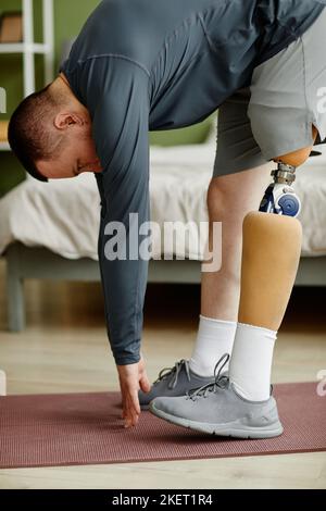 Vertikales Porträt eines Mannes mit einem prothetischen Bein, der zu Hause trainiert und Stretching-Übungen macht Stockfoto