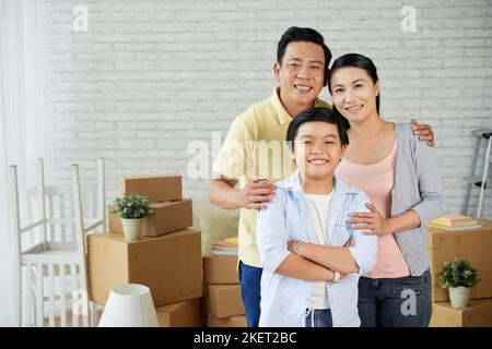 Gruppenportrait einer glücklichen asiatischen Dreiergruppe, die mit einem breiten Lächeln auf die Kamera blickt, während sie vom Auspacken bewegter Boxen abgelenkt wird, und der Inneneinrichtung einer neuen Wohnung im Hintergrund Stockfoto