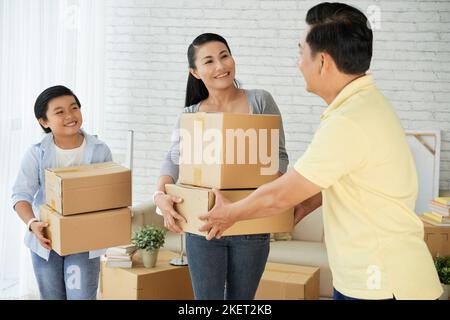 Attraktive asiatische Frau, die Stapel von Umzugskartons an ihren Mann übergibt, während sie in Packsachen eingewickelt ist, um in eine neue Wohnung umzuziehen. Ihr kleiner Sohn hilft ihr dabei Stockfoto