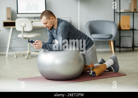 Seitenansicht Porträt eines erwachsenen Mannes mit Beinprothese, der zu Hause und mit dem Smartphone mit Fitnessball trainiert Stockfoto