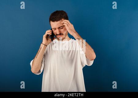 Kaukasischer junger Mann trägt T-Shirt posiert isoliert auf blauem Hintergrund Gefühl verärgert verzweifelt reden am Telefon Probleme. Hört sich schlecht neu an Stockfoto