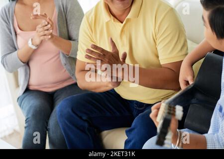 Nahaufnahme eines nicht erkennbaren Jungen, der mit Begeisterung Gitarre spielt, während seine Eltern auf dem Sofa sitzen und ihm applaudieren Stockfoto