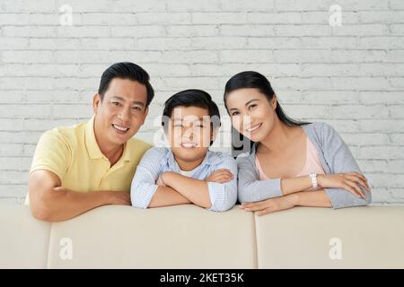 Fröhliche asiatische Familie, die sich auf dem gemütlichen Sofa lehnt und mit einem breiten Lächeln auf die Kamera schaut, während sie den Sonntag zu Hause gemeinsam verbringt, Gruppenporträt Stockfoto