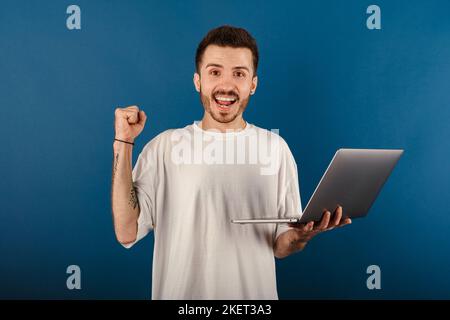 Kaukasischer Mann mit weißem T-Shirt, der isoliert auf blauem Hintergrund mit einem Laptop-Computer posiert und dabei eine Gewinnergeste macht. Stockfoto