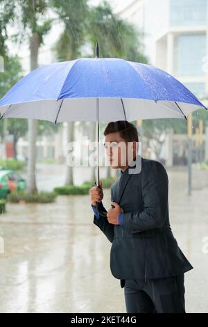 Porträtaufnahme eines gutaussehenden vietnamesischen Geschäftsmannes in klassischem Anzug, der wegschaut, während er im Freien mit Regenschirm in der Hand im strömenden Regen steht Stockfoto