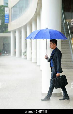 Profilansicht eines selbstbewussten asiatischen Managers, der einen klassischen Anzug trägt und einen Regenschirm in den Händen hält, während er im Freien bei strömendem Regen läuft Stockfoto