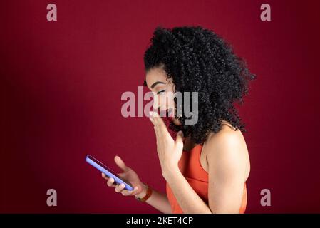 Junge, schöne, fröhliche Frau, die ihr Handy anschaut, in orangefarbener Kleidung. Isoliert auf rotem Studiohintergrund. Stockfoto