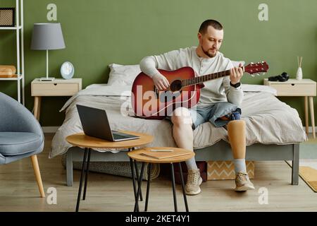 Ganzkörperportrait eines Mannes mit Beinprothese, der zu Hause Gitarre spielt und einen Laptop verwendet, Kopierraum Stockfoto