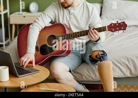 Nahaufnahme eines Mannes mit Beinprothese, der zu Hause Gitarre spielt und einen Laptop benutzt, Kopierraum Stockfoto