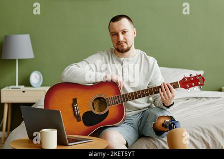 Porträt eines Mannes mit Beinprothese, der zu Hause Gitarre spielt und einen Laptop benutzt Stockfoto