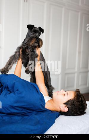 Glücklich lächelnder Junge auf dem Bett liegend und lachend in der Kamera mit seinem Hund. Konzept von glücklichen Kindern, die gute Zeit zu Hause haben. Stockfoto