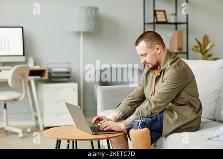 Seitenansicht Porträt eines Mannes mit Beinprothese mit Laptop, während er in minimalem Büroinnenraum arbeitet, Kopieplatz Stockfoto